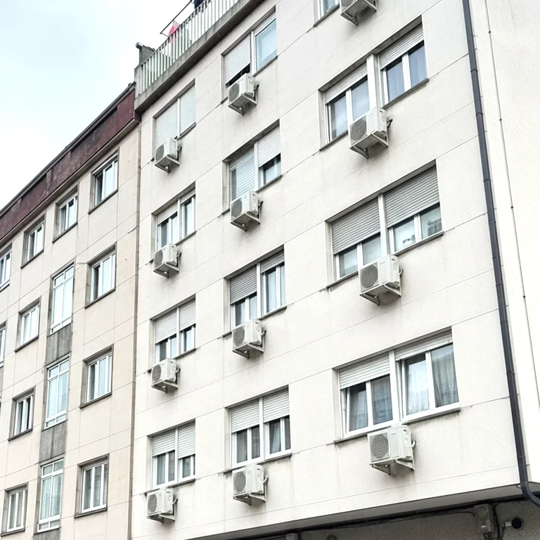 Fachada blanca de cuatro plantas con múltiples unidades exteriores de aire acondicionado expuestas en las ventanas.